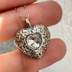 May include: A heart-shaped pendant necklace featuring a textured, dark metallic finish with a faceted, clear heart-shaped stone set in the center. The pendant hangs from a thin, silver-toned metal chain with small, uniform circular links.