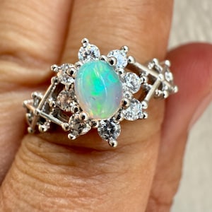 May include: A silver-toned ring featuring a central oval-shaped opal with iridescent green and blue flashes. The opal is surrounded by a halo of small, round, clear faceted stones, with additional clear stones set along the split metal band.