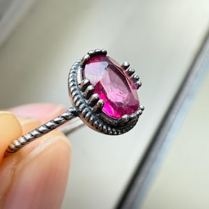 May include: A close-up of a silver ring featuring a large, oval-cut, magenta gemstone. The ring has a textured band and a decorative bezel setting around the stone. The ring is held by a person's fingers.