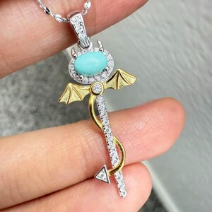 May include: A silver necklace with a pendant featuring a turquoise oval stone surrounded by small clear stones. The pendant has gold-colored wings and a silver-colored staff with clear stones. The pendant is in the shape of a key.