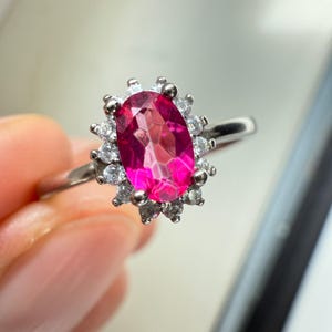 May include: A silver ring with a striking, oval-cut, hot pink gemstone at its centre, encircled by a halo of small, clear, round-cut stones. The ring has a thin, polished band, and the setting is a classic, elegant design.