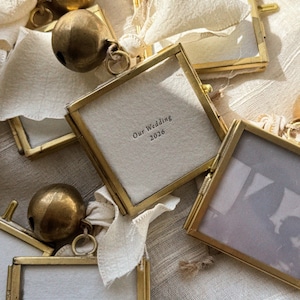 May include: Square brass-framed glass ornaments featuring off-white fabric inserts with printed text reading Our Wedding 2026 and Our First Wedding Anniversary 2026, each accented with a large round antique brass bell and a cream-colored fabric ribbon.