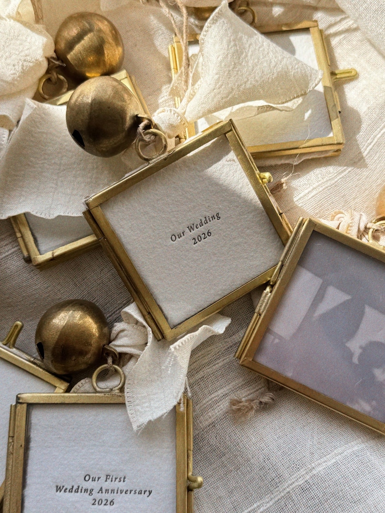 May include: Square brass-framed glass ornaments featuring off-white fabric inserts with printed text reading Our Wedding 2026 and Our First Wedding Anniversary 2026, each accented with a large round antique brass bell and a cream-colored fabric ribbon.