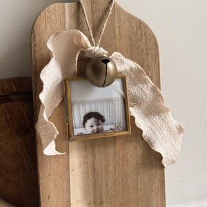 May include: A square brass picture frame containing a photograph of a child, suspended by a jute string from a wooden cutting board. A large, round, matte brass bell and a long, textured, cream-colored fabric ribbon are tied to the top of the frame.