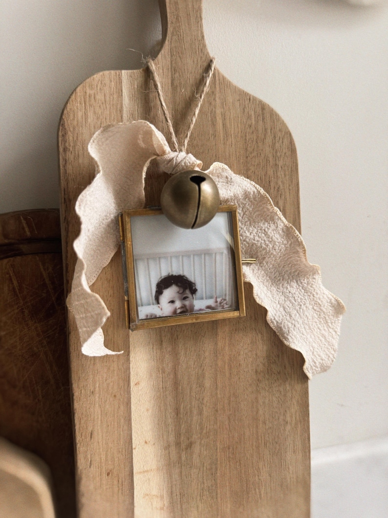 May include: A square brass picture frame containing a photograph of a child, suspended by a jute string from a wooden cutting board. A large, round, matte brass bell and a long, textured, cream-colored fabric ribbon are tied to the top of the frame.
