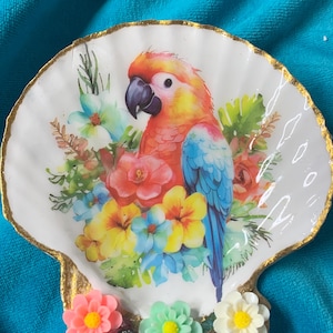 May include: A decorative seashell-shaped trinket dish with a gold-painted rim. The dish features a colorful parrot illustration surrounded by flowers in shades of red, yellow, and blue. Three small, colorful flower embellishments are attached to the base.