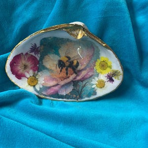 May include: A decorative seashell with a gold-painted rim. The shell interior features a floral design with a bumblebee, surrounded by dried flowers in shades of pink, yellow, and white. The shell rests on a turquoise fabric.