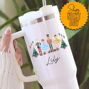 May include: White travel mug with a handle, featuring a colorful illustration of characters from The Nutcracker ballet, including a mouse, a ballerina, and a toy soldier. The name "Lily" is written in cursive below the illustration. The mug has a white lid and a straw.