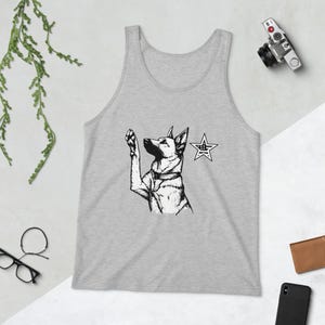 May include: A grey vest top with a black and white illustration of a dog raising its paw, and a star graphic. The dog is wearing a collar. A camera, wallet, and phone are in the background.