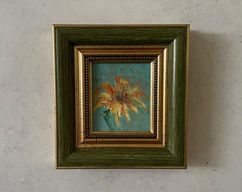 May include: A framed painting of a yellow flower with orange accents on a blue background. The frame is green with gold accents.