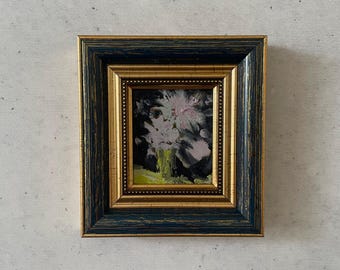 May include: Small framed floral oil painting. The artwork features a bouquet of white and pink flowers in a green vase against a dark background. The frame is a combination of gold and navy blue.
