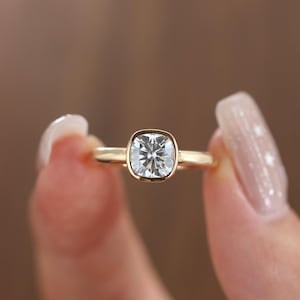 May include: A gold ring featuring a square-cut, clear gemstone. The ring is held between two fingers against a blurred brown backdrop. The gemstone is set in a bezel setting, and the band is thin and polished.