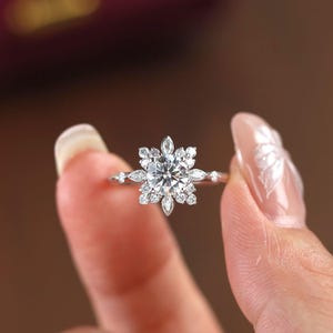 Puede incluir: Un anillo plateado con una piedra central redonda y transparente, rodeada de piedras más pequeñas y transparentes dispuestas en un diseño floral o de copo de nieve. El anillo es sostenido por dedos, mostrando los intrincados detalles de la joya.