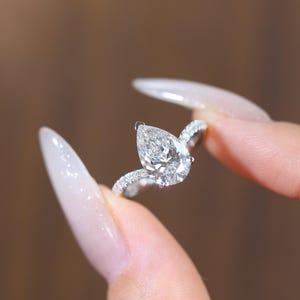 May include: A close-up of a diamond ring with a pear-shaped center stone. The ring is set in a silver band with smaller diamonds. The ring is held between two fingers against a blurred brown background.
