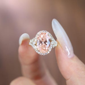 Op de afbeelding: Close-up van een ring met een grote, ovaal geslepen perzik-kleurige edelsteen, omringd door een halo van kleinere, heldere stenen. De ring heeft een delicate, sierlijke band met bladachtige details, in een gouden tint.