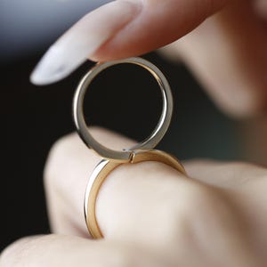 May include: Close-up of two gold rings, one held above the other. The rings are simple bands with a polished finish. The focus is on the rings, with a blurred background.