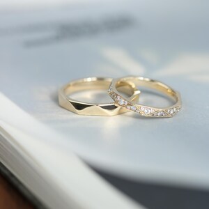 May include: Two gold wedding rings. One ring features a geometric pattern, and the other is embellished with small, sparkling stones. The rings are set against a soft, blurred background, highlighting their intricate details and elegant design.