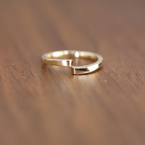 May include: A gold-colored ring with a unique, modern design. The ring features a split band with a slight overlap, creating an open, geometric shape. The ring is set against a warm, brown wooden surface.