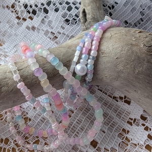 May include: A set of pastel-coloured beaded bracelets with a single pearl accent. The bracelets are layered on a piece of driftwood.