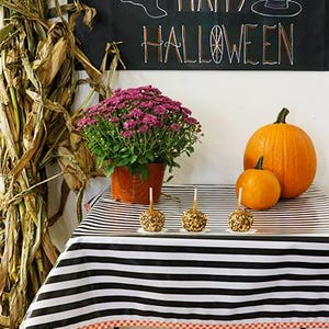 May include: A Halloween-themed display featuring a black and white striped tablecloth with caramel apples, pumpkins, and a potted plant. A banner reads "HAPPY HALLOWEEN" with a ghost and witch hat.