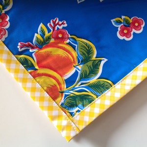 May include: A corner of a blue tablecloth with a vintage fruit and floral pattern. The design features large orange peaches, red flowers, and green leaves. The edge is trimmed with a yellow and white gingham ribbon.