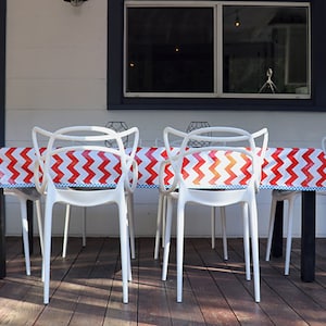 May include: A patio dining set featuring a rectangular table with a red and white chevron patterned tablecloth. Six white plastic chairs surround the table. The table has black legs and is set on a wooden deck.
