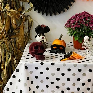 May include: A Halloween-themed display featuring a red velvet skull, a pumpkin with a black mask, and a white tablecloth with black and gray polka dots. Decorations include a skeleton, a black paper fan, and a pot of purple flowers.