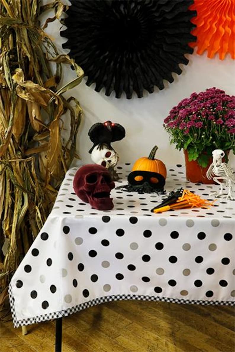 May include: A Halloween-themed display featuring a red velvet skull, a pumpkin with a black mask, and a white tablecloth with black and gray polka dots. Decorations include a skeleton, a black paper fan, and a pot of purple flowers.