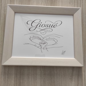 Puede incluir: Un cuadro enmarcado con un marco de madera clara. La obra de arte presenta un dibujo lineal de manos formando un corazón alrededor de la mano de un bebé. El nombre "Giosue" está escrito arriba, con la fecha 14/06/25 debajo.
