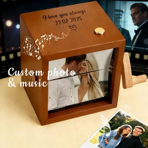 以下が含まれることがあります： キスをしているカップルの写真が入った、茶色の木製オルゴール。「I love you always 22.07.2025」と「Custom photo & music」の文字が入っています。上部には金のノブがあり、側面には音符が描かれています。