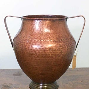 Puede incluir: Un jarrón de cobre martillado con dos asas curvas. El jarrón tiene un cuerpo ancho y redondeado y una banda decorativa cerca de la parte superior. Se asienta sobre una base circular escalonada. Una pequeña etiqueta cuelga del lateral. El jarrón mide unos 30 cm de altura.