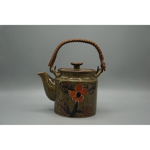 May include: A ceramic teapot with a brown and orange floral design. The teapot has a woven handle and a lid. The body of the teapot is a speckled brown color. The floral design features an orange flower with brown leaves.