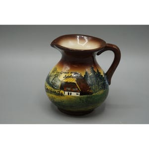 May include: A ceramic pitcher with a brown glazed rim and handle, featuring a hand-painted landscape scene of a house with a thatched roof, green trees, and a grassy field. The text "Grüß Gott Schwarzwald" is written above the house.