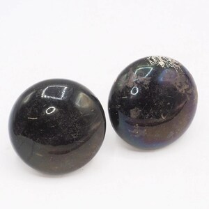 May include: Two smooth, spherical objects with a dark, polished finish. The surfaces are primarily black with subtle iridescent purple and blue undertones visible under light. Each sphere features a glossy, reflective texture with minor surface variations.