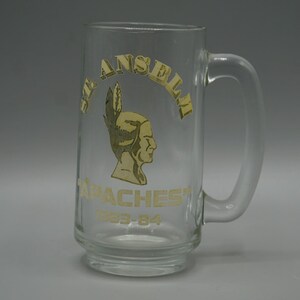 May include: A clear glass beer mug featuring a gold-colored graphic of a person wearing a feathered headdress. The text reads ST. ANSELM, APACHES, and 1983-84 in gold lettering. The mug has a sturdy handle and a thick, reinforced base design.