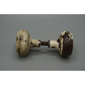 May include: An antique door knob with a distressed, aged appearance. The knob features a dark brown central rod connecting two rounded, off-white knobs with visible wear and chipping, suggesting age and use. The hardware is likely made of metal.