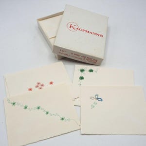 May include: A vintage Kaufmann's stationery set in a white box labeled 12 Deckle Edge Notes and 12 Envelopes. The set includes cream-colored note cards with deckle edges, featuring small, delicate floral illustrations in red, green, and blue ink.