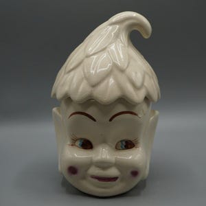 May include: A vintage ceramic jar in the shape of a smiling elf face. The jar has a cream-colored finish with brown eyebrows, blue eyes, and pink cheeks. The lid is shaped like a leafy hat.