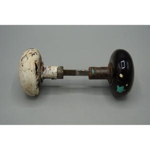 May include: An antique door knob with a white, chipped porcelain handle on one end and a black, chipped porcelain handle on the other. The connecting rod is metal with signs of rust and wear.