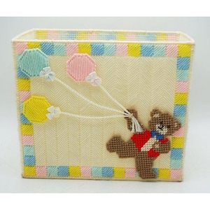 May include: A rectangular plastic canvas storage box featuring a cream-colored herringbone pattern body with a border of alternating pink, yellow, and blue squares. A brown teddy bear wearing a red vest holds three pastel-colored balloons.