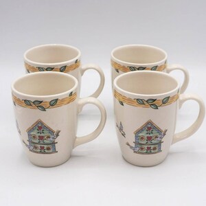 May include: Set of four ceramic mugs with a birdhouse and birds design. The mugs are white with a yellow and green band around the top. The birdhouse is blue and green with red hearts. The birds are blue and white.