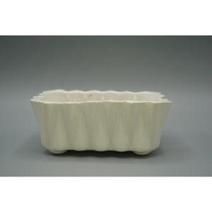 May include: A rectangular off-white ceramic planter featuring a vertical fluted or pleated texture along the exterior walls. The vessel has a scalloped top edge and is supported by small, integrated feet at each corner of the base.