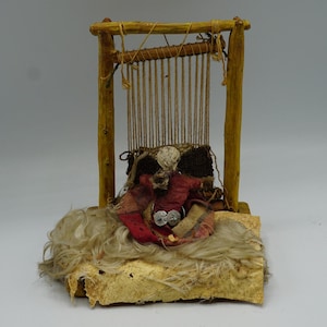 May include: A miniature wooden loom with a woven textile in shades of red, brown, and cream. The loom is constructed from light-colored wood and features vertical threads. The textile incorporates silver-colored embellishments and rests on a textured base.