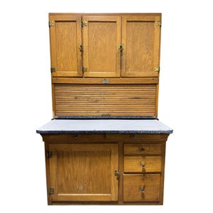 May include: An antique wooden kitchen cabinet with a light brown finish. The cabinet features upper storage with three doors, a roll-top section, and a lower section with a door and drawers. The countertop has a speckled, light gray surface.