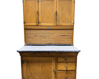 Könnte beinhalten: Ein antiker Küchenschrank aus Holz mit hellbraunem Finish. Der Schrank verfügt über obere Aufbewahrung mit drei Türen, einen Rollladenabschnitt und einen unteren Abschnitt mit einer Tür und Schubladen. Die Arbeitsplatte hat eine gesprenkelte, hellgraue Oberfläche.