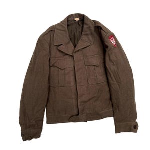 May include: A brown military jacket with two chest pockets and two lower pockets. The jacket has a button closure and a small emblem on the upper left sleeve. The jacket is made of a wool-like material.