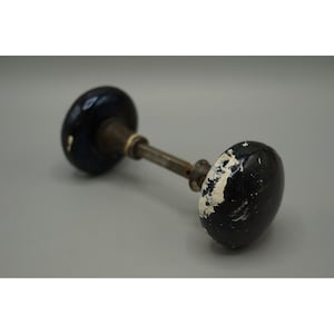 May include: An antique door knob with a dark blue, oval-shaped handle and a tarnished metal rod. The handle has white paint chipping off, revealing the dark blue underneath. The door knob is set against a neutral background.