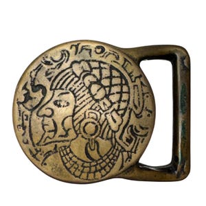 May include: A circular brass belt buckle featuring an embossed profile of a figure wearing an ornate headdress and a wide collar necklace. The design includes intricate line work and geometric patterns surrounding the central portrait figure.