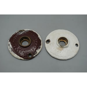 May include: Two antique door knob backplates. One is painted white, the other is painted dark red with white chipping paint. Both have a central hole and two smaller holes for screws.