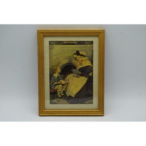 May include: A framed print featuring a vintage illustration of a person in a bonnet and dark dress sewing while sitting near a fireplace, with a child in light-colored clothing sitting on a small stool nearby reading a book in a gold frame.